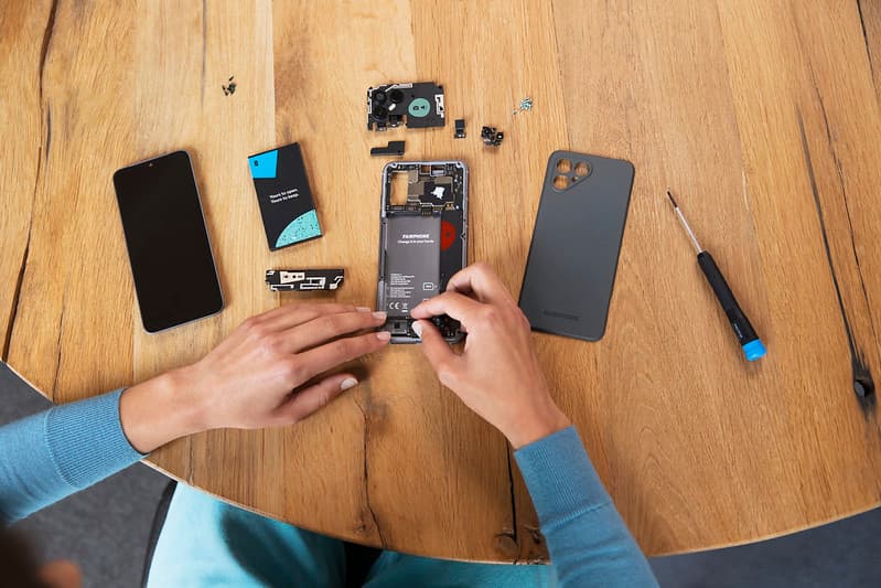 A phone is laid out in parts on a wooden table. A screwdriver with a blue and black handle is visible on the right. Two hands are busy repairing the phone.