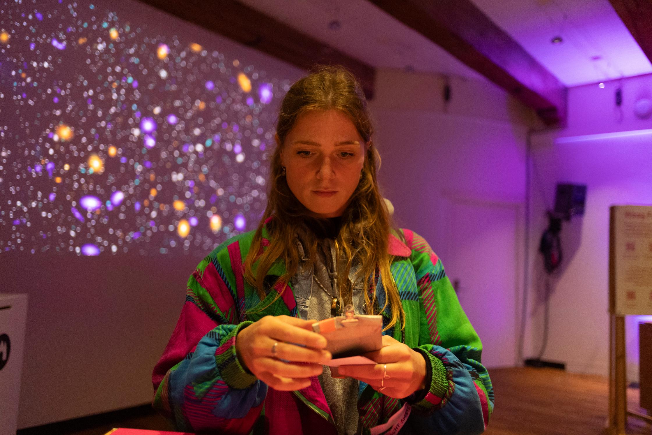 Een vrouw met een velgekleurd jasje houd een apparaat vast. Achter haar is een sterrenhemel geprojecteerd tegen een muur met gele en paarse sterren.