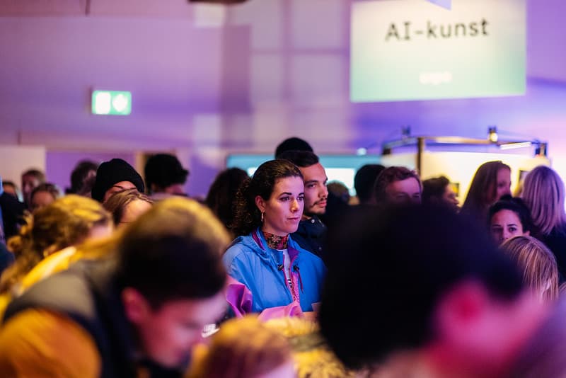 De foto toont een druk publiek tijdens museumnacht in Waag. Een persoon met een blauwe jas is scherp in beeld in het midden, ze kijkt naar een beeldscherm. Om haar heen zie je mensen die onscherp in beeld staan. Op de achtergrond zie je een uithangbord met de tekst AI-kunst, ook onscherp.