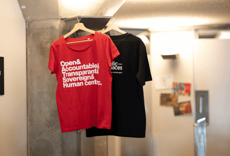 Een rood shirt hangt aan een haakje met de woorden Open & Accountable & Transparent & Souvereign & Human centric. Er hangt ook een wart t-shirt achter met het public spaces logo. 