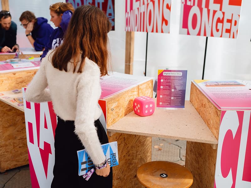 A woman is standing in front of a wooden counter. On the counter lies a large pink die and there is a sign with instructions for Digital Detox Dice.