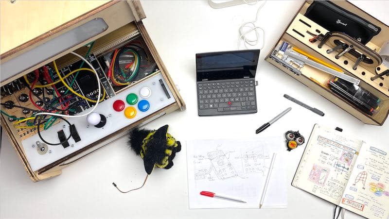 Picture of the work in progress of the Furby synthesiser module by Edwin Dertien. On the left there is a large wooden box opened with various electronic elements, such as big colorfol buttons, wires, switches and lights. Next to it there is a small laptop and a deconstructed Furby toy. There is also a technical drawing of the device  and on the right a wooden toolbox like box with pens and other tools. 