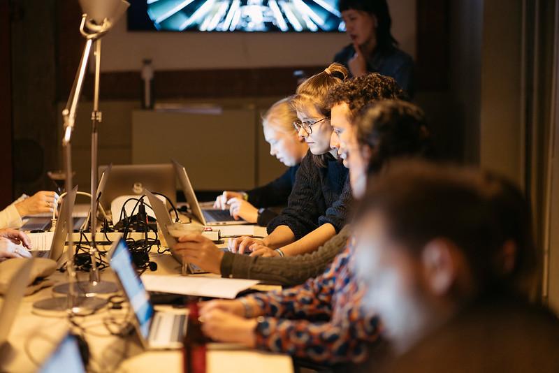 Aan een lange tafel zitten meerder mensen achter laptops te werken. 