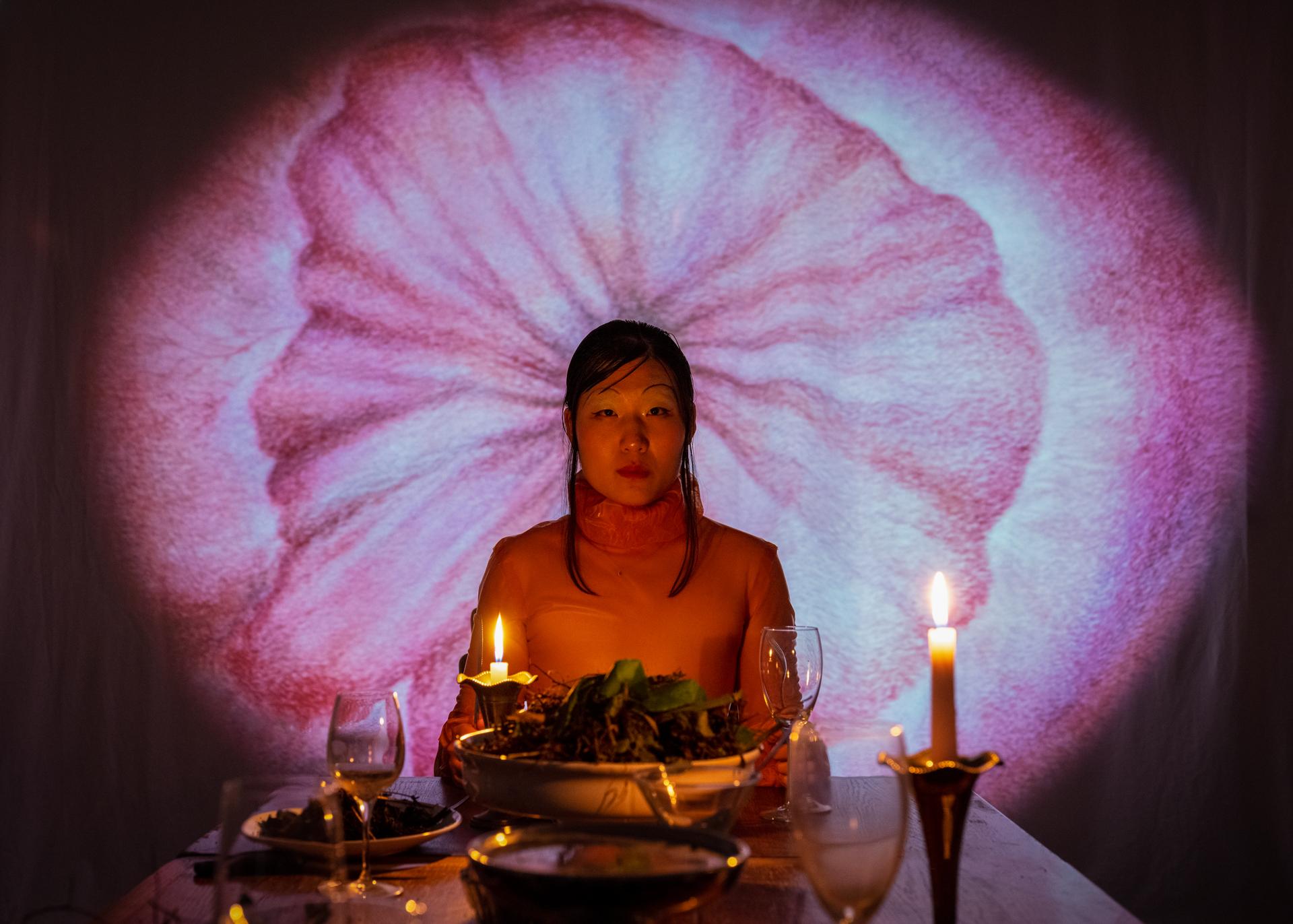 Portrait photo of Kexin Hao, sitting at a table with two candles besides her and a large pink/purple image of a flower behind her head.