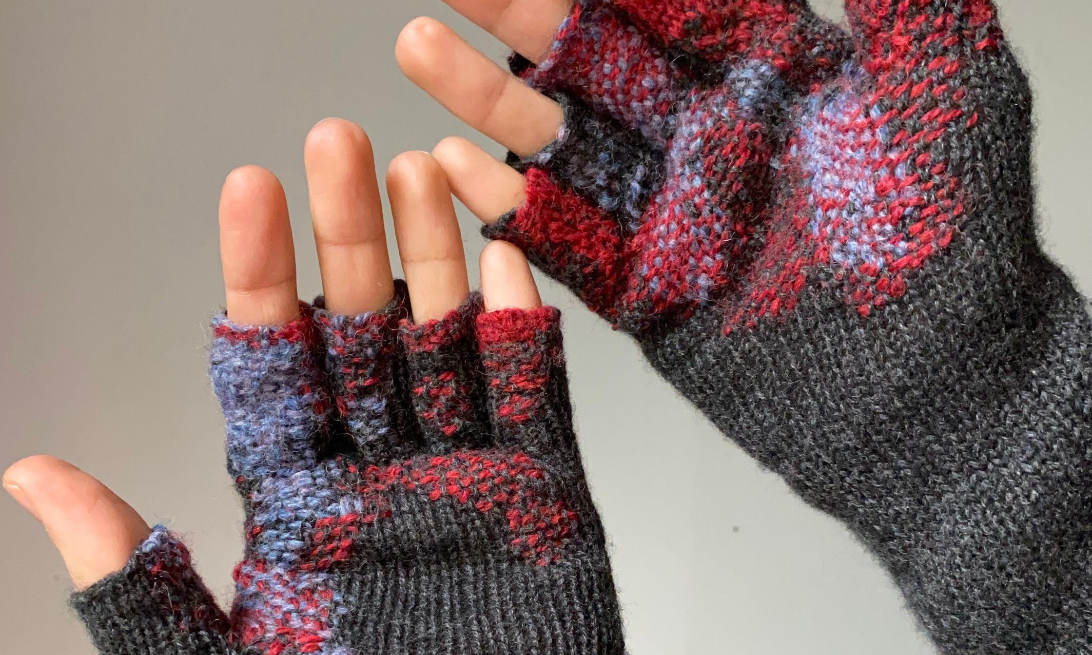 Two hands wearing dark grey fingerless gloves are visible, their palms facing towards the camera. The gloves have been mended with intricate blue and red patterns.