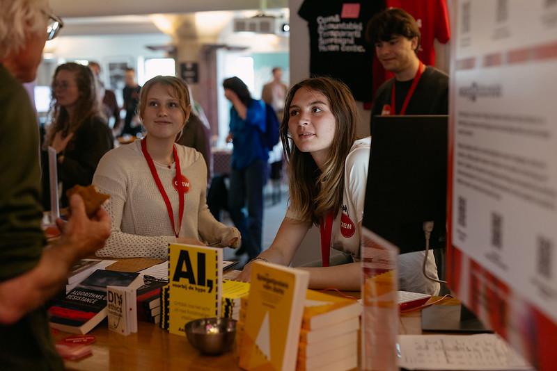 Twee vrouwen zitten aan een tafel en een bezoeker spreekt ze toe. Op de tafel zijn boeken uitgestald, eentje over A.I..