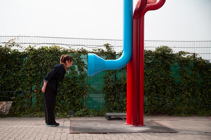Een vrouw bukt naar voren om te kunnen kijken in een grote blauwe kijkbuis die in een hoek naar boven loopt. Een andere rode buis gaat recht naar beneden.