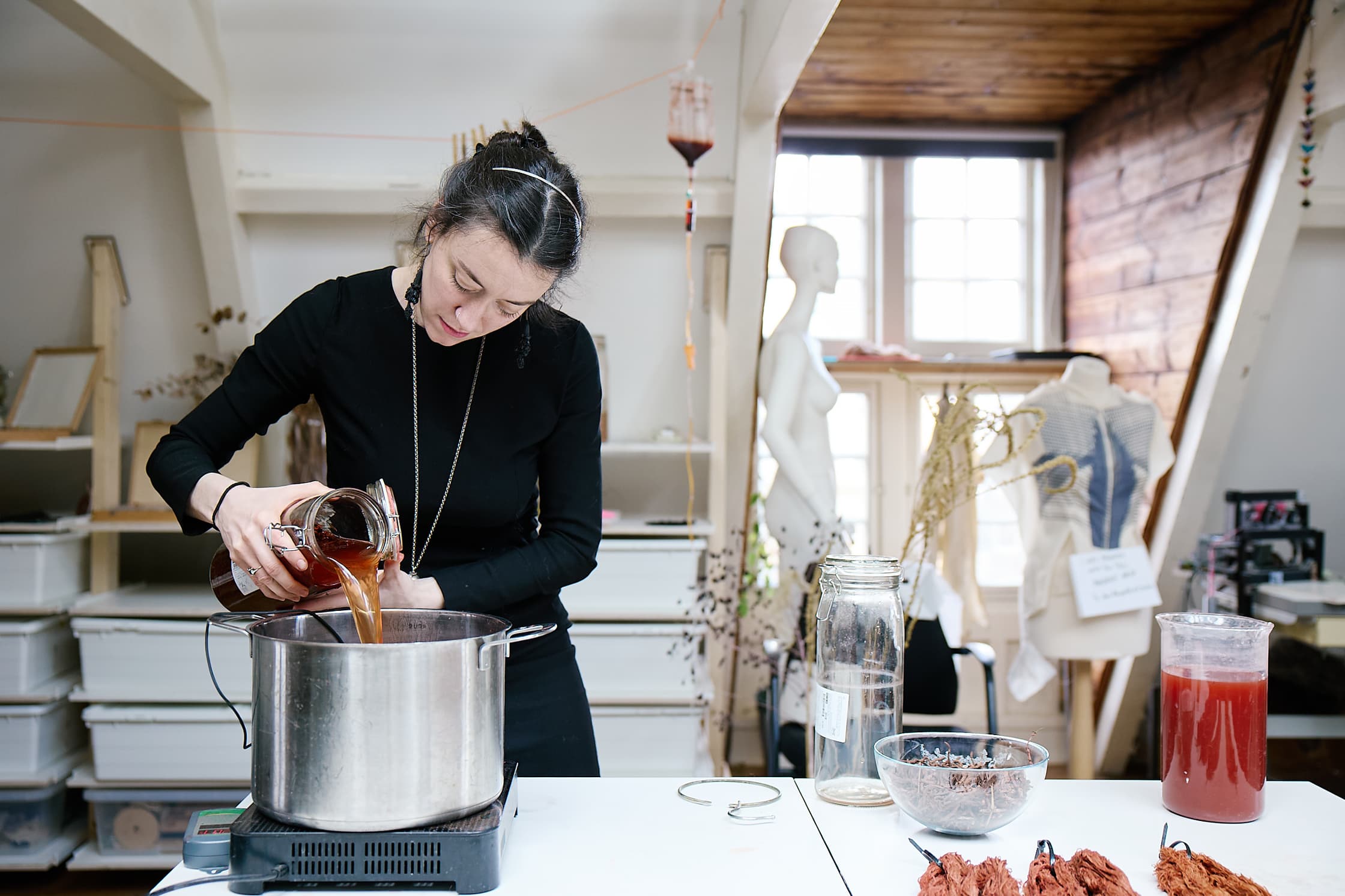 Foto van het TextileLab in de Waag, een zolderachtige ogeving. Een vrouw met zwart haar giet een brion-oranje vloeistof in een grote metalen pan. Op de achtergrond zien we een aantal kasten en mannequins met verschillende vloeistoffen en textielen.