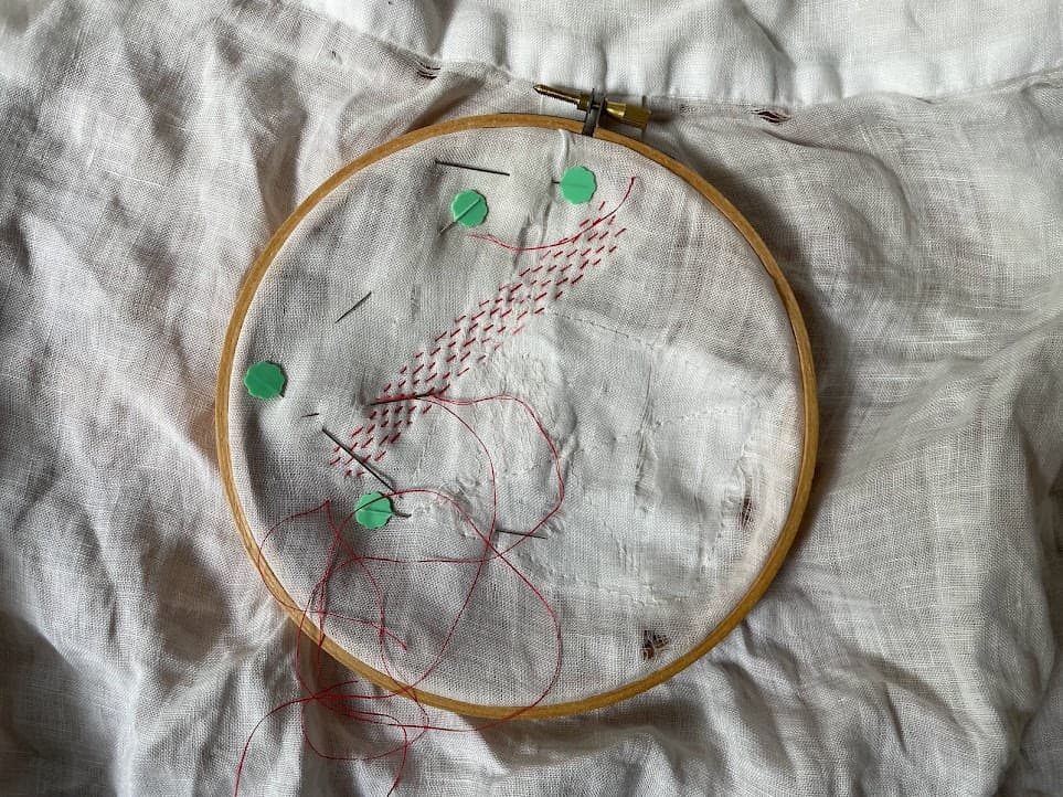 A piece of white fabric is covered with a wooden stitching ring. There are stitches visible, existing of little squares of mint green yarn