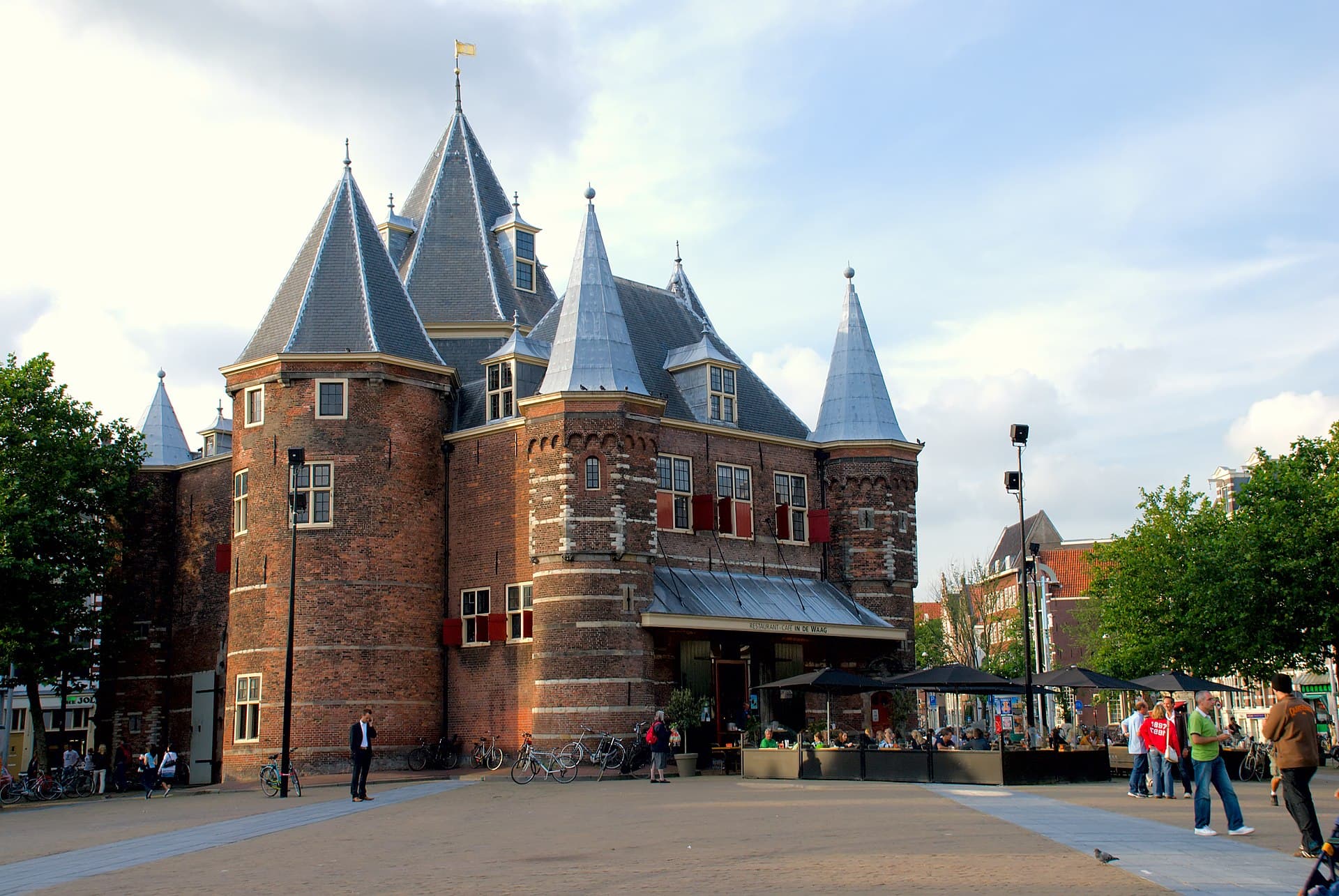 Foto van het Waag gebouw in Amsterdam aan de Nieuwmarkt. De Waag is een oude stenen stadspoort met meerdere torens. Op de begane grond is een terras te zien van het restaurant In de Waag.
