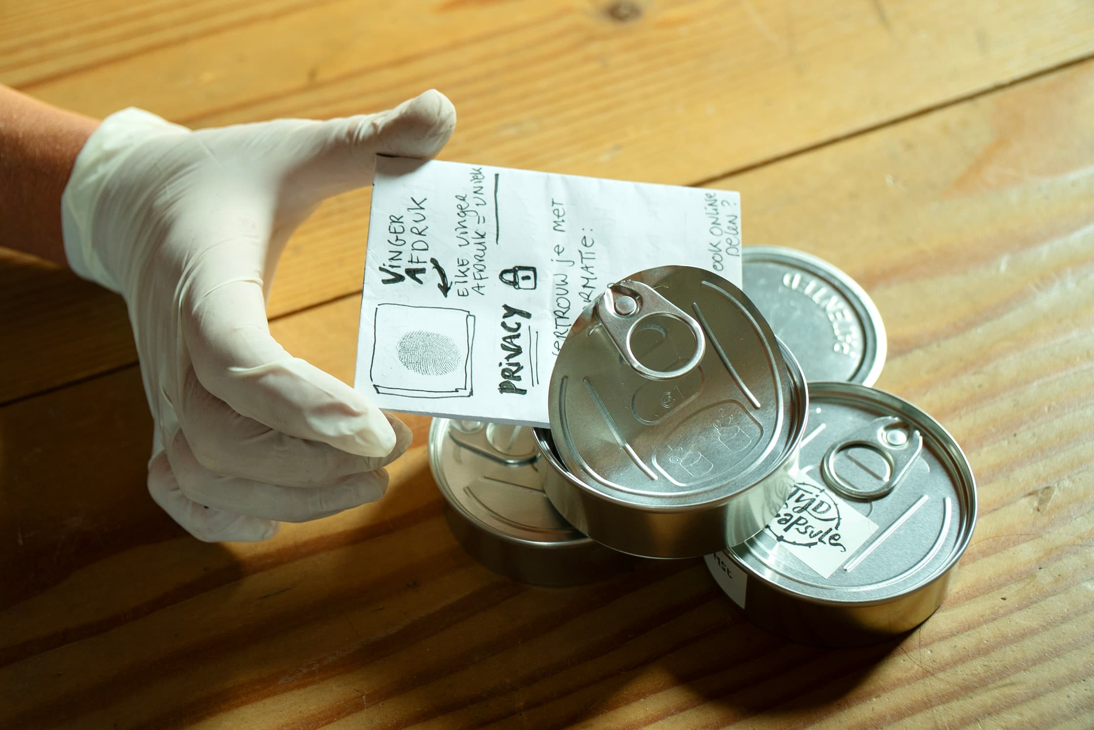 Foto van een houten tafelblad waarop drie kleine ronde blikjes liggen. Een hand met witte latex handschoen houd een papiertje vast waarop staat 'Vinger afdruk, elke afdruk is uniek' en het woordje 'privacy' met een slotje. In een vierkantje staat een vingerafdruk stempel. De hand stopt het papiertje in een van de blikjes.