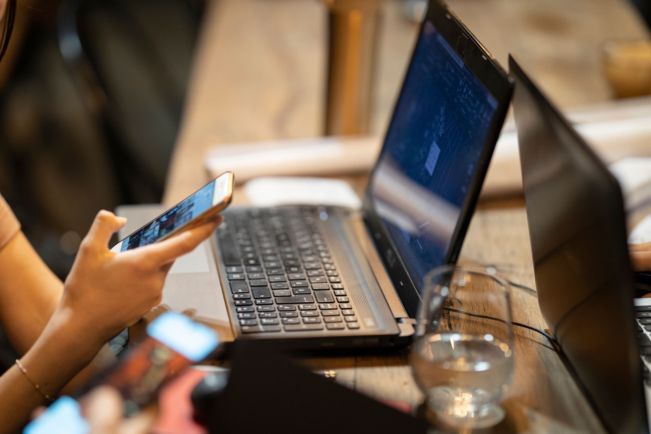 Een open laptop op tafel en iemand die erachter zit met telefoon in zijn hand.
