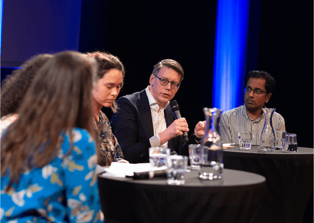 Foto tijdens een panelgesprek in de OBA. Vier mensen zitten aan ronde tafels met zwarte bekleding, voor hun staan glazen met water.