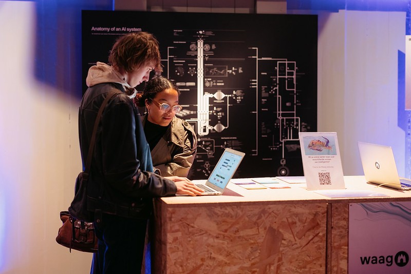 Twee mensen staan aan een houten tafel en kijken naar een laptop. Achter hen hangt een zwarte plaat aan de muur met daarop de tekst 'The anatomy of an AI system'.