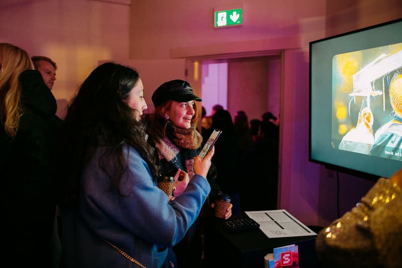 Op de voorgrond zie je twee mensen die kijken naar een scherm. Een vrouw met zwarte krullen houdt een telefoon vast. Een persoon naast haar met baseball petje kijkt naar het scherm en praat.