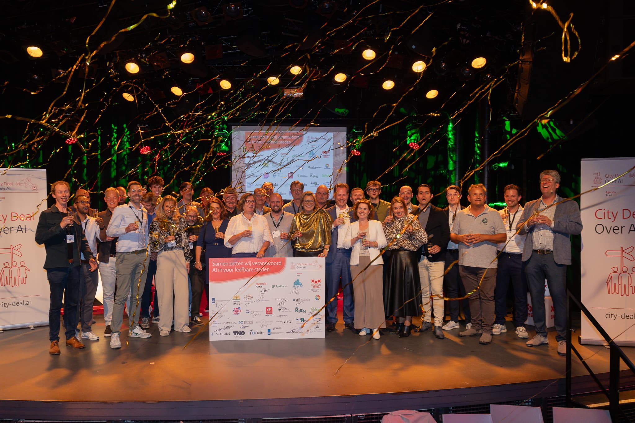 Groepsfoto genomen tijdens de ondertekening van de City Deal over AI. Een groep mensen staat op een podium en houden een glas champagne omhoog. Goude lintjes dwarrelen door de lucht. Op een groot bord zijn logo's van verschillende organisaties te zien met handtekeningen met de tekst: Samen zetten wij verantwoord AI in voor leefbare steden.
