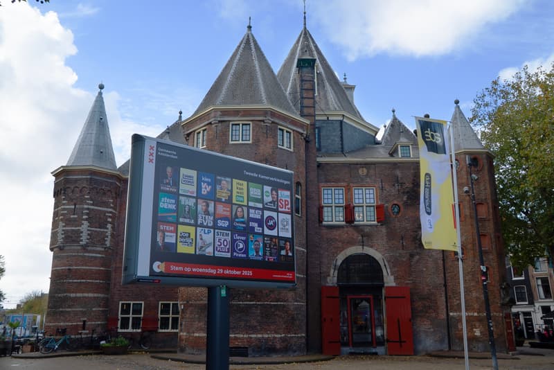 Foto van een verkiezingsbord met alle partijposters (trotter) voor het gebouw van de Waag op de Nieuwmarkt.