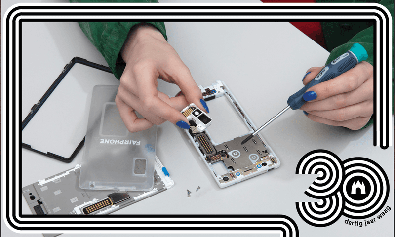 Een Fairphone ligt in verschillende onderdelen uiteen op een wit tafelblad. Twee handen zijn in beeld die een chip vasthouden en deze in de telefoon leggen.