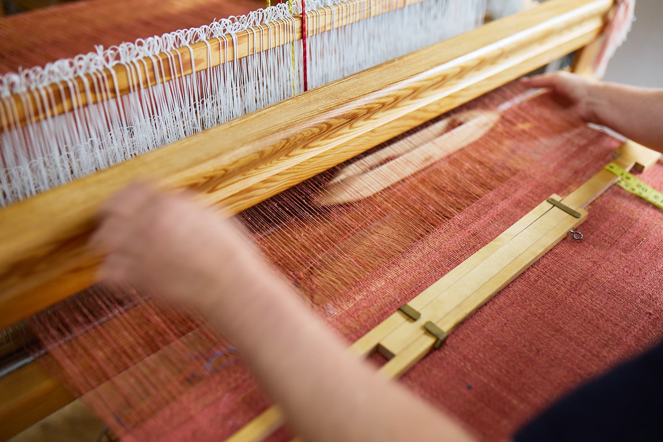 Een foto van een houten weefgetouw: een houten frame waarop linnen draden zijn gespannen. Het linnen dat gespannen is over het getouw heeft een roodoranje, terracotta achtige kleur. Twee handen bedienen het weefgetouw.