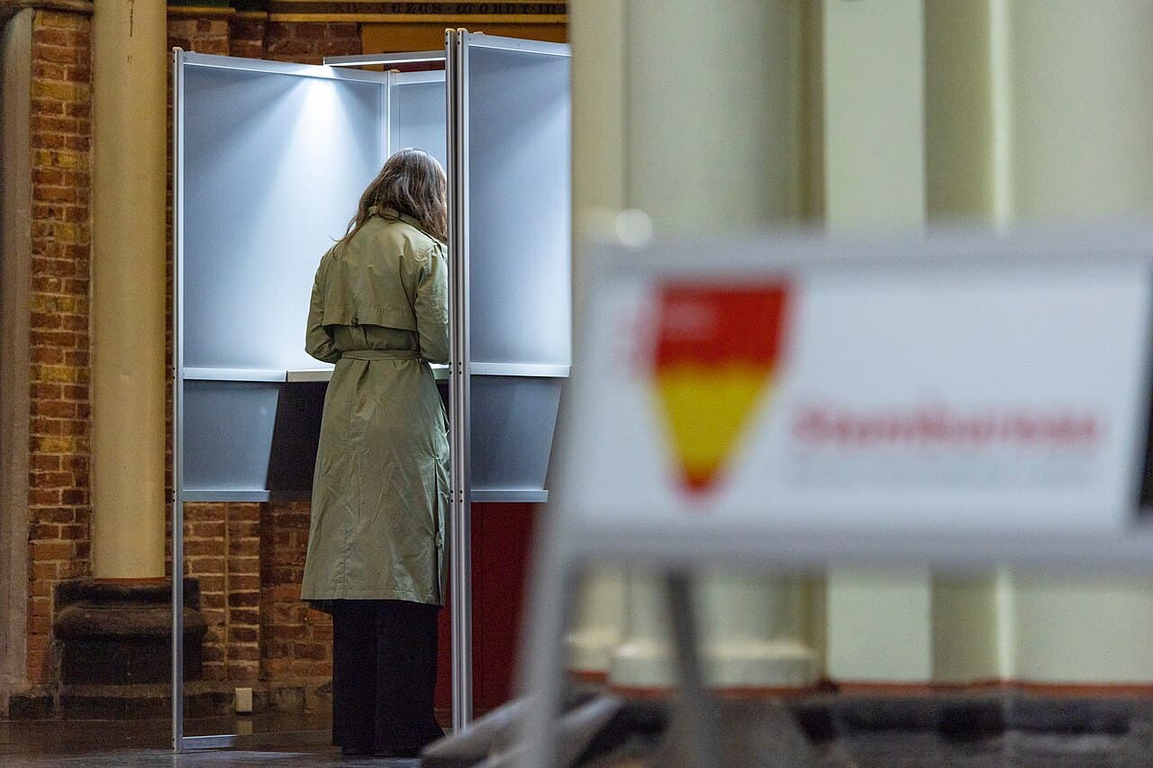 Een persoon met een beige regenjas staat in een stemhokje, rechts ervan een bord met rood potlood en de tekst Stemlocatie.