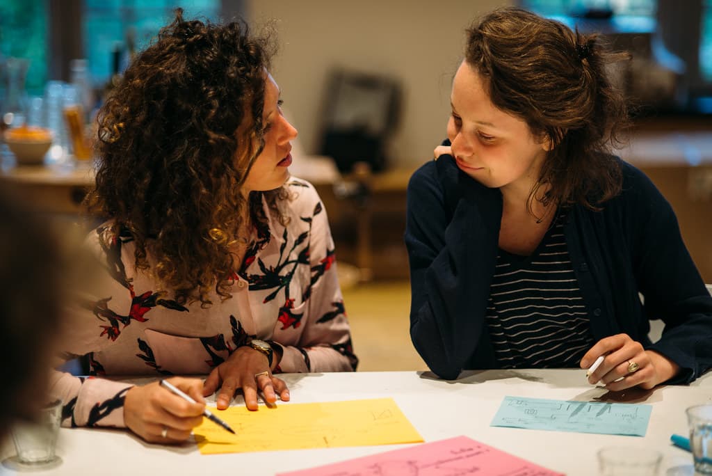 Foto van twee mensen aan tafel in gesprek met elkaar, voor hun op tafel liggen kleurige papiertjes.