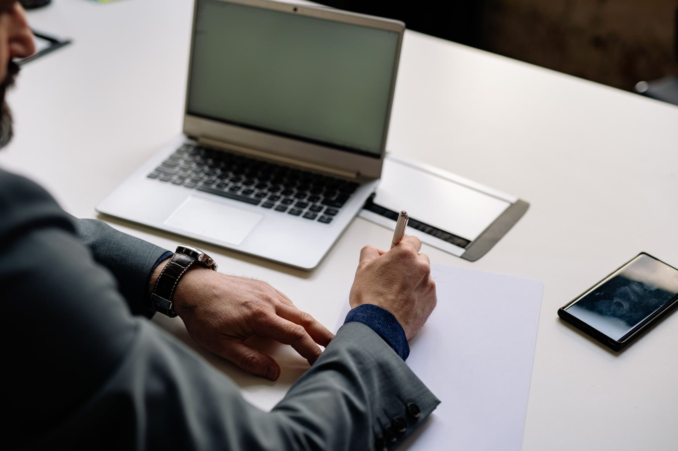 A man in a suit writing on a piece of paper with his laptop and phone on his desk