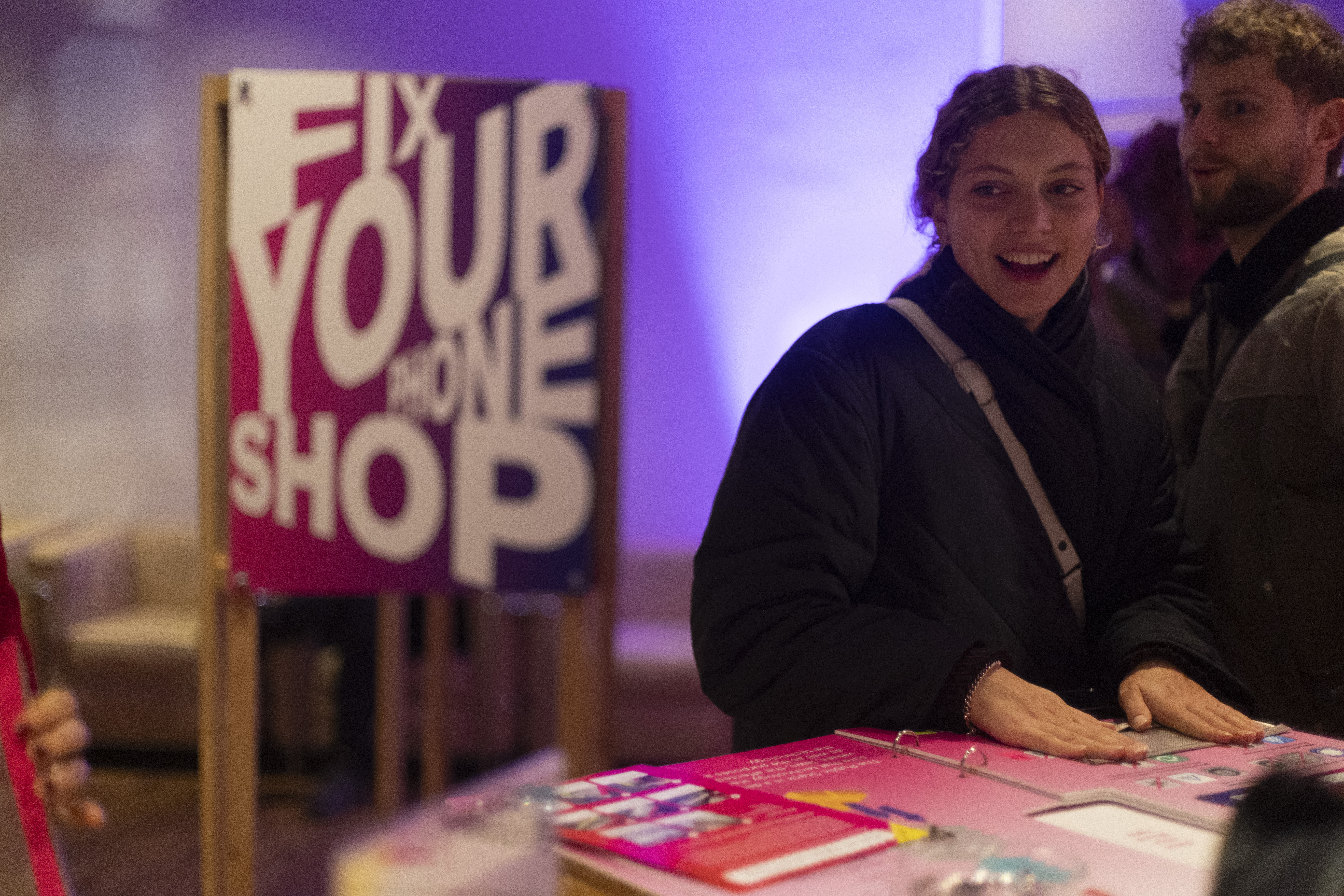 Een vrouw staat aan een tafel met daarop felgekleurde paarse flyers. Ze kijkt opzij. Achter haar staat een houten bord met 'Fix your phone shop' in grote witte letters tegen een rood-paarse achtergrond.