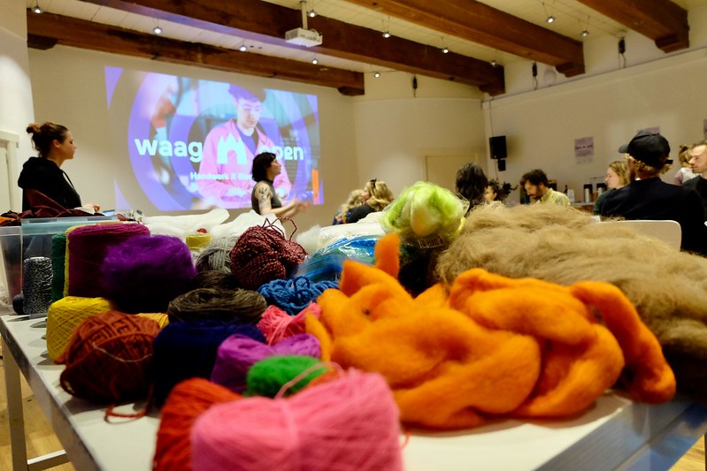 Photo with colorful yarns and fabrics layed out on a table. In the background there is someone presenting a talk with the Waag Futurelab logo projected on a wall behind them.