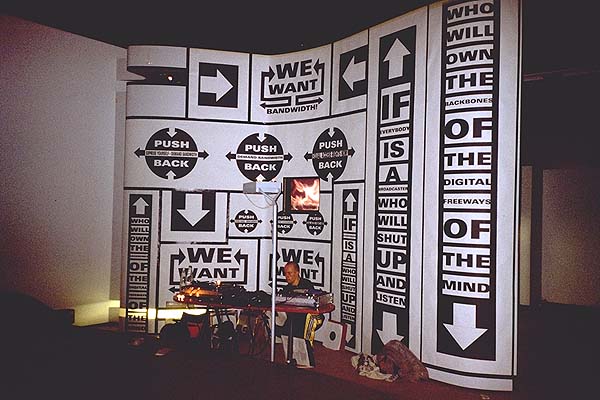 Foto van de We want bandwidth manifestatie tijdens Hybrid Workspace in Kessler, 1997. Een man zit achter een tafel met computerapperatuur. Achterhem zijn hoge wanden te zien met grote zwarte iconen van pijlen en cirkels met teksten als 'We ant bandwidth' en 'push back'.
