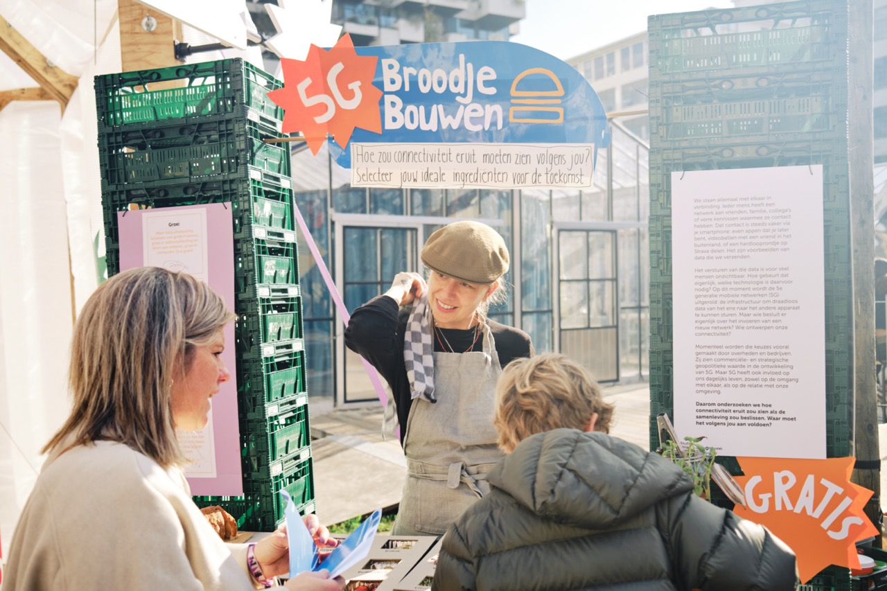 Een toonbank opgebouwd uit groene kratjs. Boven de toonbank hangt een bord met '5G broodje bouwen. Hoe zou connectiviteit eruit moeten zien volgens jou? Selecteer jouw ideale ingredienten voor de toekomst'. Achter de toonbank staat een vrouw met een theedoek over haar schouder en een schort. Ze helpt een vrouw met een kind met een broodje.