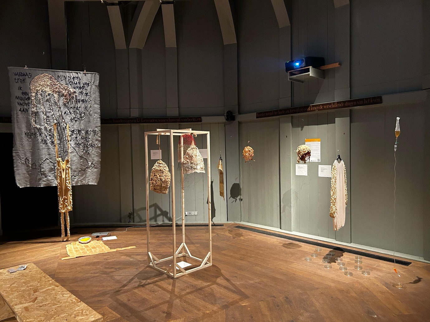 Installation view of the Fabricademy show, a dark hexagonical shaped space with sage green wooden walls is lit by a few small lights, and several pieces of fabric are displayed throughout the room.