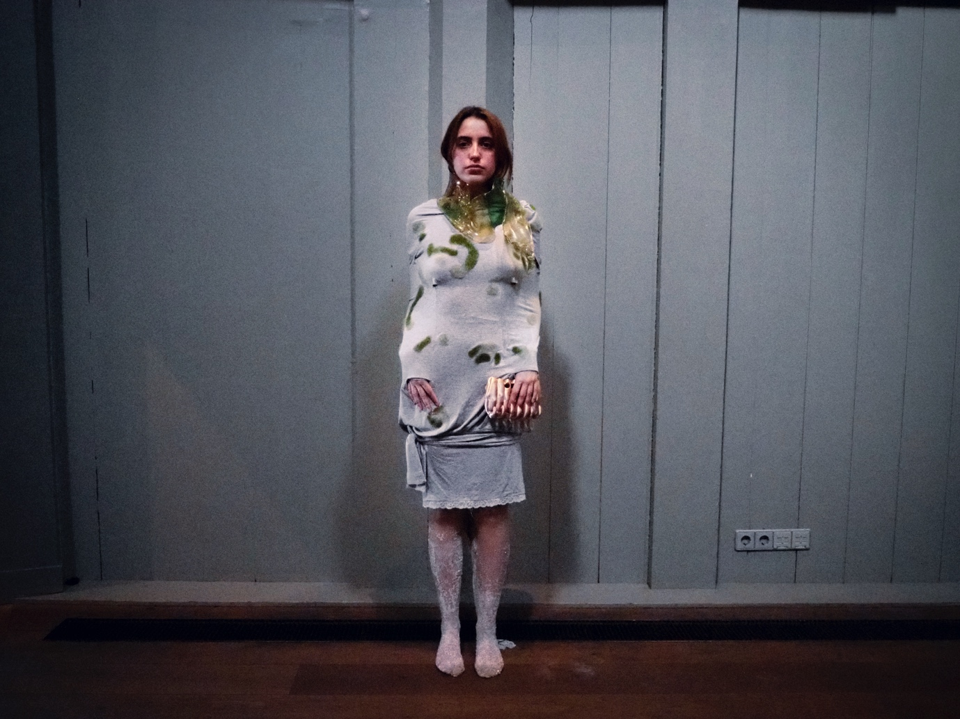 A woman is standing with her back to the wall in a dimly lit room, wearing a striped scarf with gelatine bio-silicone, which bears a resemblance to bubbly lake water. She is also wearing abamboo jersey dress with needle-felted wool and a pair of crystallised tights with alum crystallisation and in her hands she is holding a wearable sculpture with 3D-printed PLA silk, which has the shape of a pair of self-hugging/clutching hands