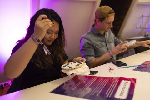 Een vrouw met lang zwart haar houd haar hand omhoog met daaraan naald en draad. Ze borduurt aan een stukje stof op een ronde borduurring. Voor haar licht een geplastificeerde tekst op een paarse achtergrond. Naast haar zit een man ook te borduren.