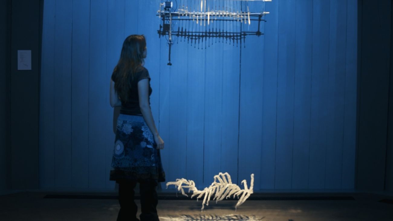 A visitor is viewing the Amphidrite installation in a dark room with dark blue lighting. There is a white skeletal structure down on the floor, and a similar, more metal structure is repeated up aboce, suspended in the air.
